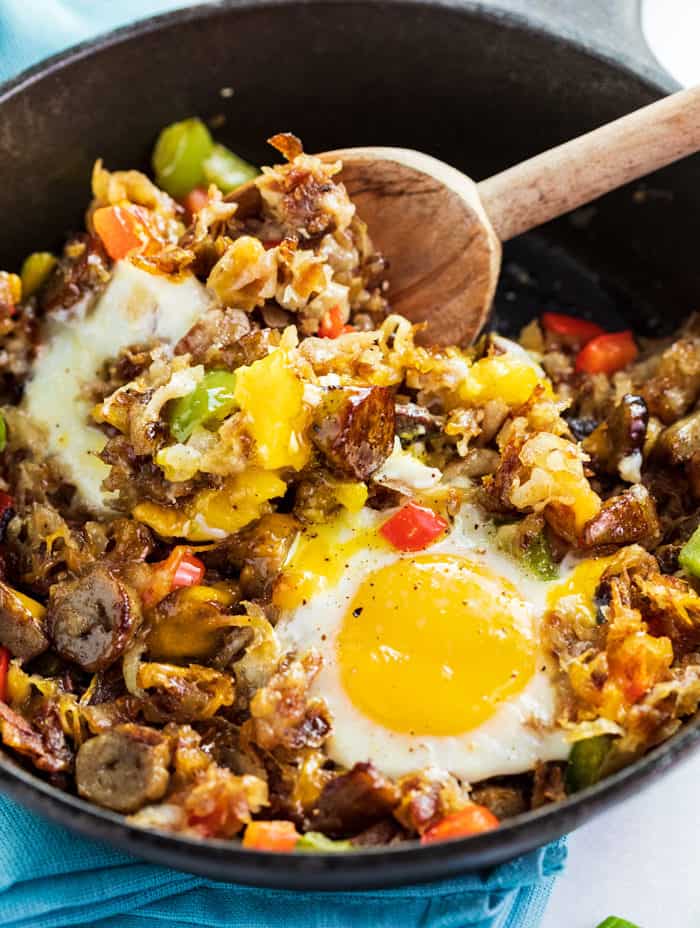 A cast iron skillet with a baked egg and a wooden spoon stirring cooked hash, sausage, and bell peppers. 