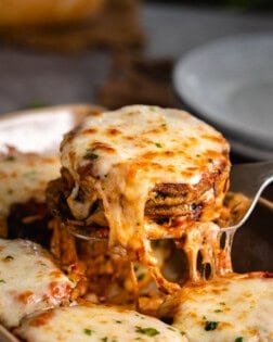 A stack of eggplant Parmesan topped with melted Mozzarella cheese being scooped up with a spatula.