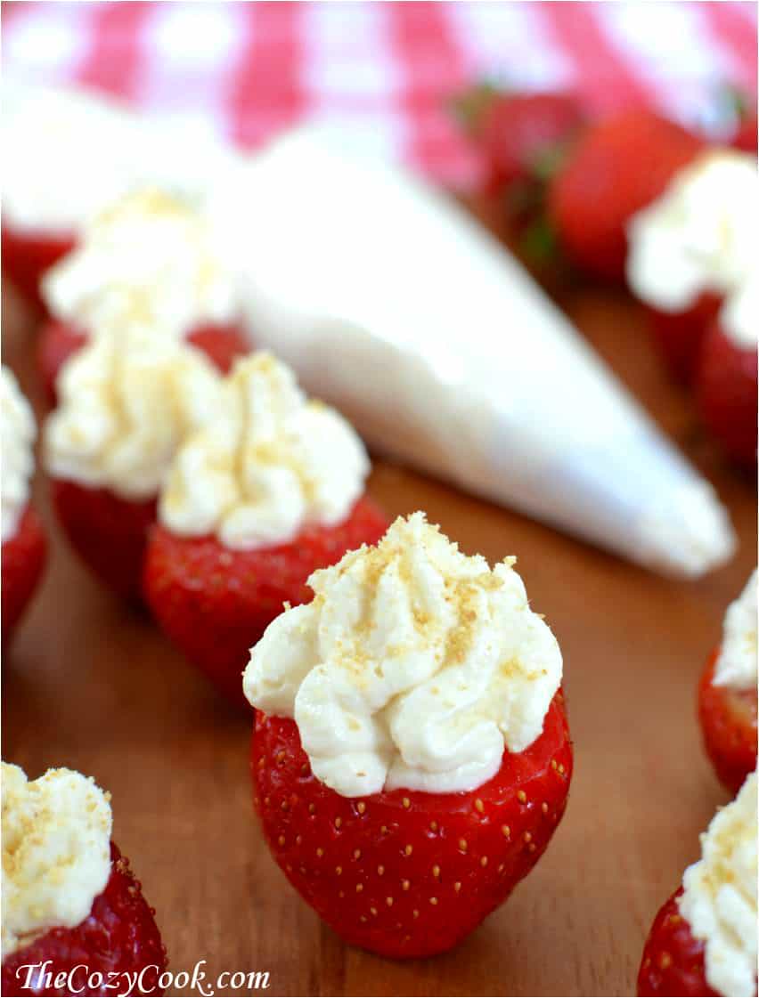 Strawberries standing on a wooden board and stuffing with cheesecake filling with white piping bag in background.