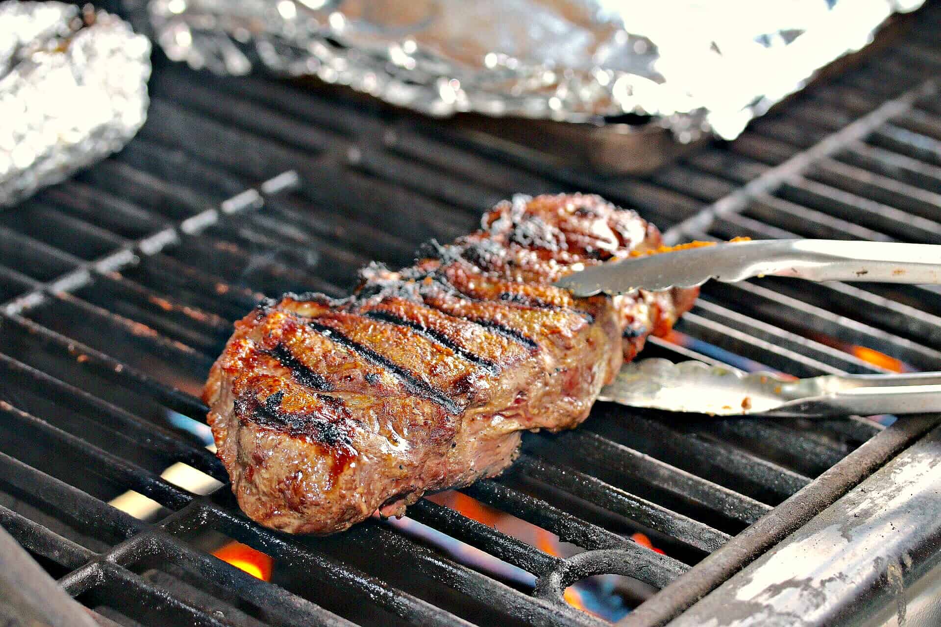 A seasoned steak on a grill with kitchen tongs lifting it up.