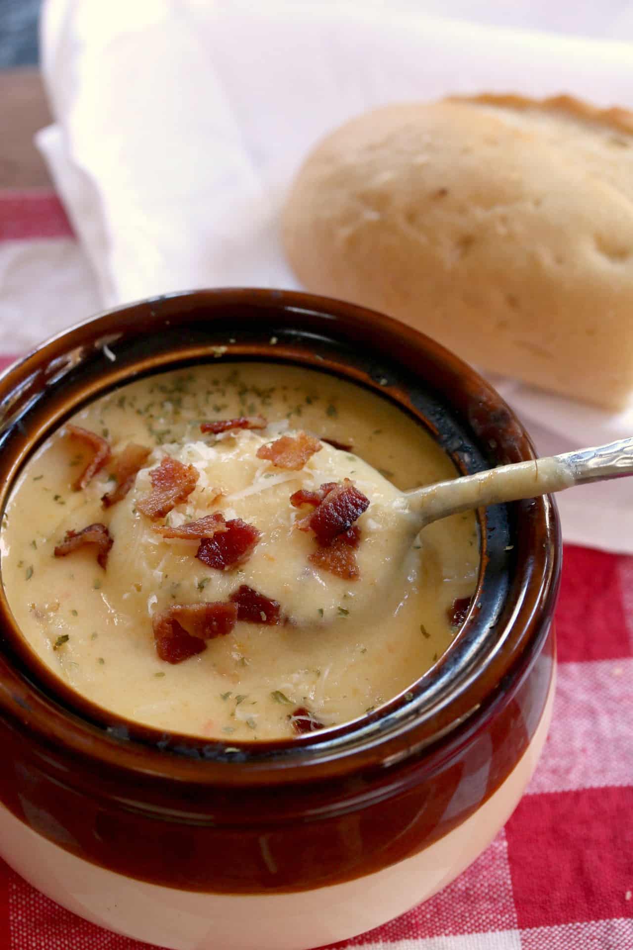 A spoon scooping up Asiago Bisque from a bowl with bacon on top and bread in the background.