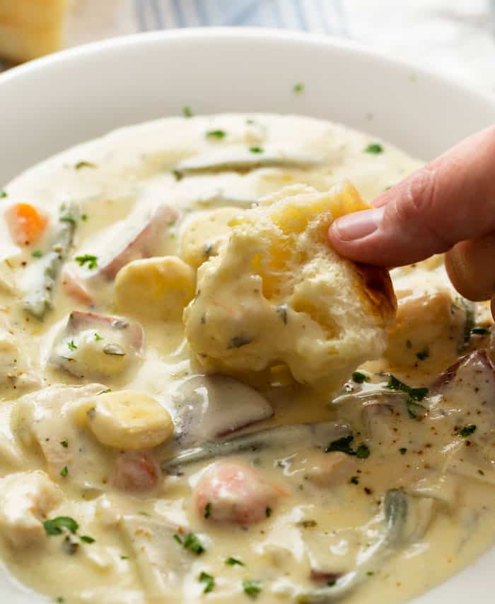 A white bowl of creamy chicken stew with hand dunking a piece of bread into the broth.