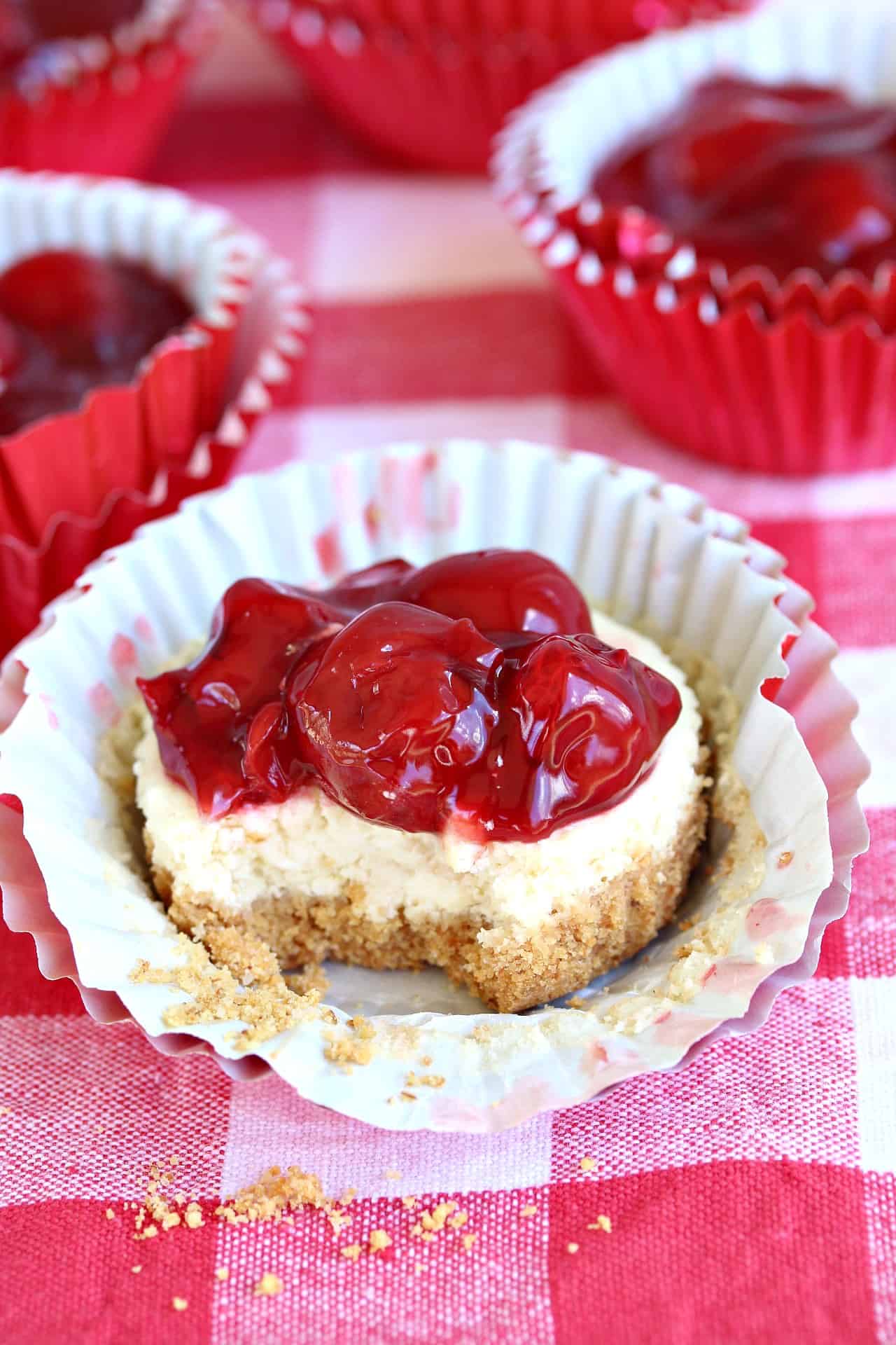 A cherry cheesecake cupcake with a bite taken out of it on a red checkered surface.