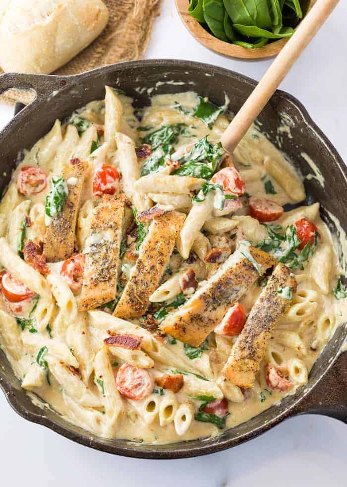 Chicken alfredo pasta in a black skillet with tomatoes and spinach.