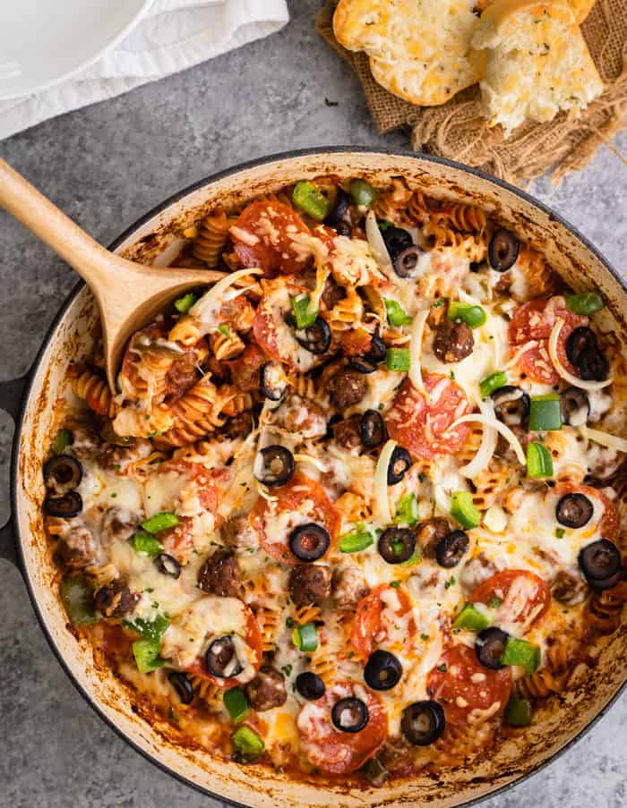 A pot of pizza pastas with a wooden spoon in it and topped with cheese, olives, pepperoni, and peppers.