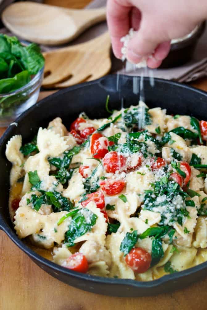 hand sprinkling parmesan cheese over skillet alfredo pasta in cast iron skillet with tomatoes and spinach