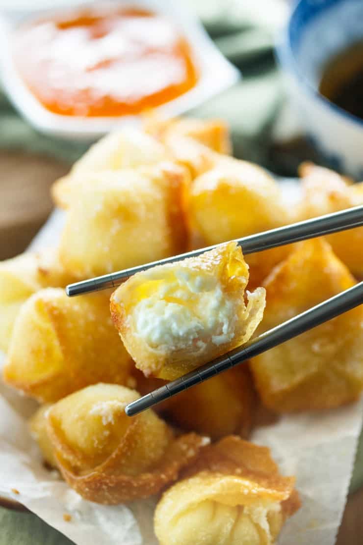 Chopsticks holding a fried wonton with cheese cheese filling inside and sweet and sour sauce in the background.
