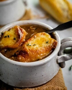 A mug of French Onion Soup topped with 2 slices of cheesy baguettes.