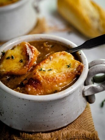 A mug of French Onion Soup topped with 2 slices of cheesy baguettes.