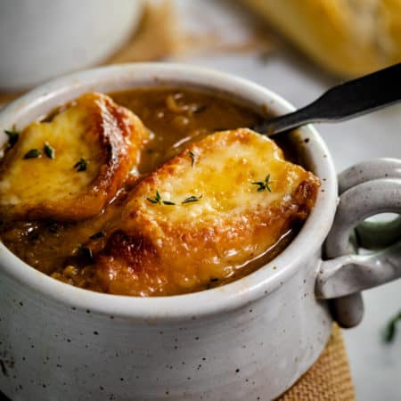 A mug of French Onion Soup topped with 2 slices of cheesy baguettes.