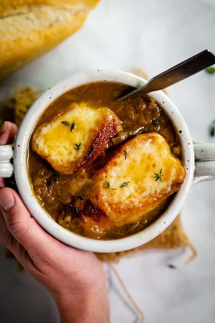 Overheat shot of a hand holding a white bowl filled with french onion soup topped with two cheesy baguettes.