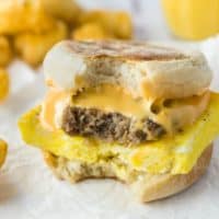 A sausage egg and cheese breakfast sandwich with tater tots and orange juice in the background.