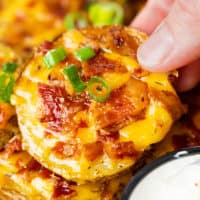 A hand holding a crispy slice of potato topped with melted cheese, bacon, and green onions.