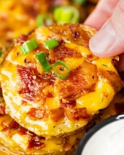 A hand holding a crispy slice of potato topped with melted cheese, bacon, and green onions.