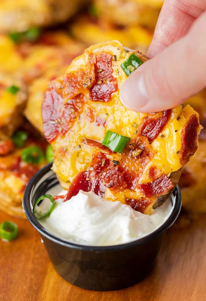 A hand dipping a baked potato slice topped with melted cheese and bacon into sour cream.