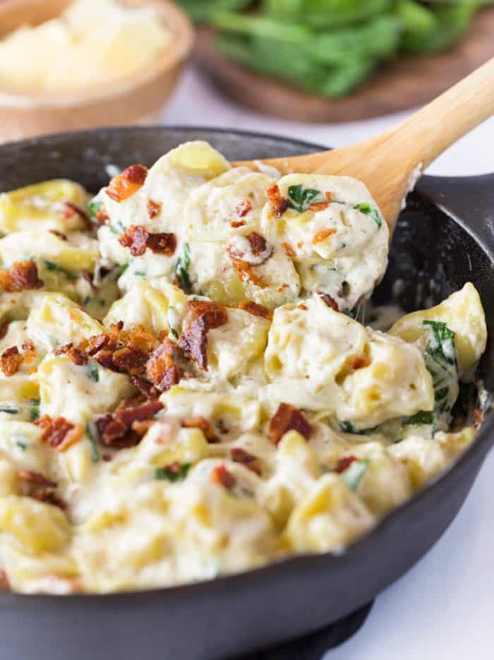 A wooden spoon scooping Tortellini Al Forno out from a cast iron skillet.