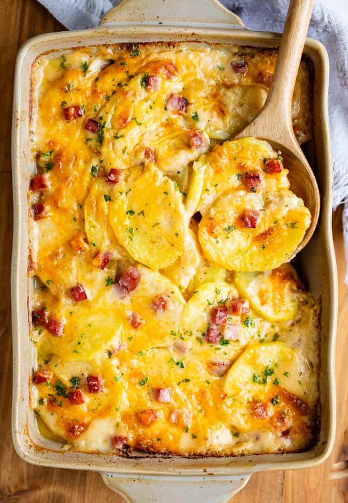 A casserole dish on wooden surface filled with Scalloped potatoes with ham with a wooden spoon in it.