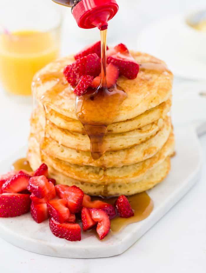 A tall stack of pancakes topped with strawberries with syrup being poured on top.