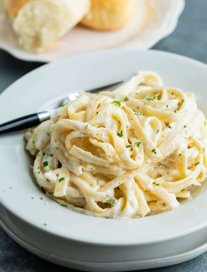 Fettuccine Alfredo sprinkled with parsley in a white bowl with rolls in the background.