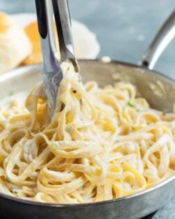 Kitchen tongs pulling up Fettuccine Alfredo from a pan.