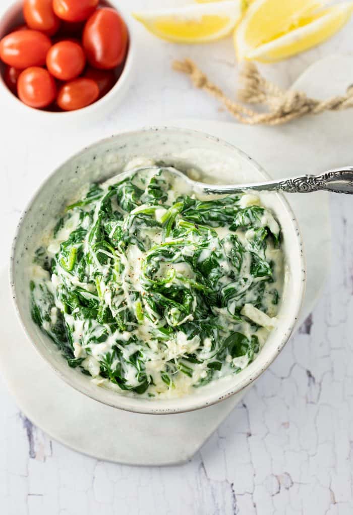 A white bowl filled with creamed spinach on a white table next to cherry tomatoes and sliced lemons.