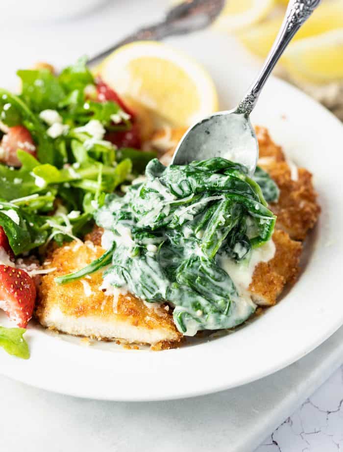 A spoon spreading creamed spinach on top of breaded chicken