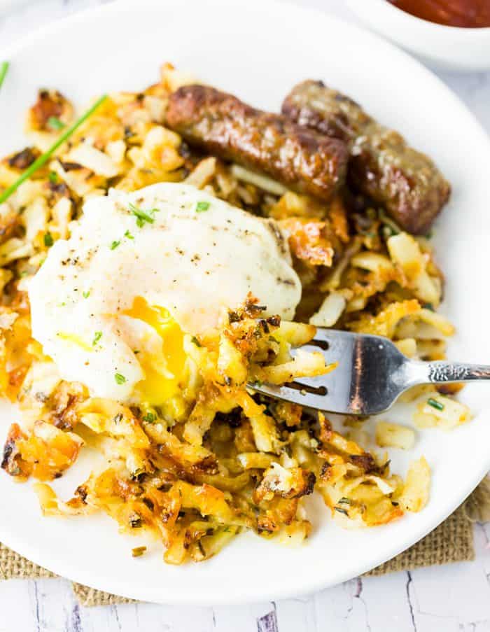 A fork scooping up waffle iron hash browns with an egg on top on a white plate.