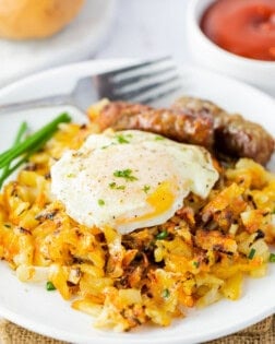 A white plate with waffle iron hash browns topped with a fried egg with sausage links in the background.