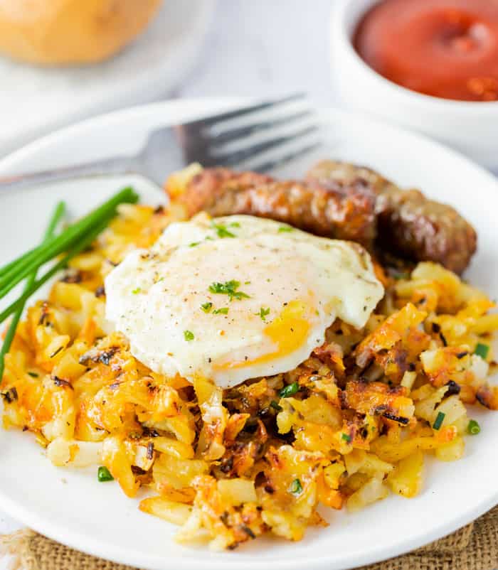 Waffle Iron Hash Browns with an egg on top on a white plate with sausage and ketchup in the background.