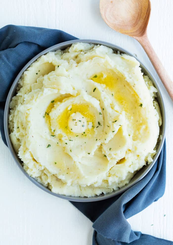 Bowl of mashed potatoes topped with melted butter on a blue dish cloth next to a wooden spoon.