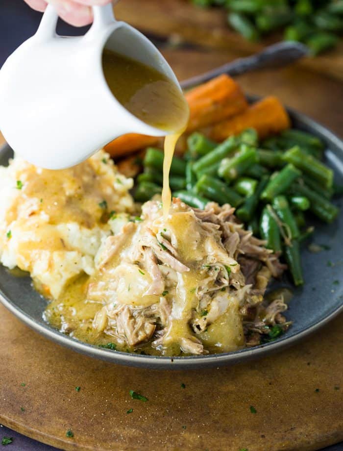 Gravy being poured over shredded pork on a plate with mashed potatoes, green beans, and carrots.