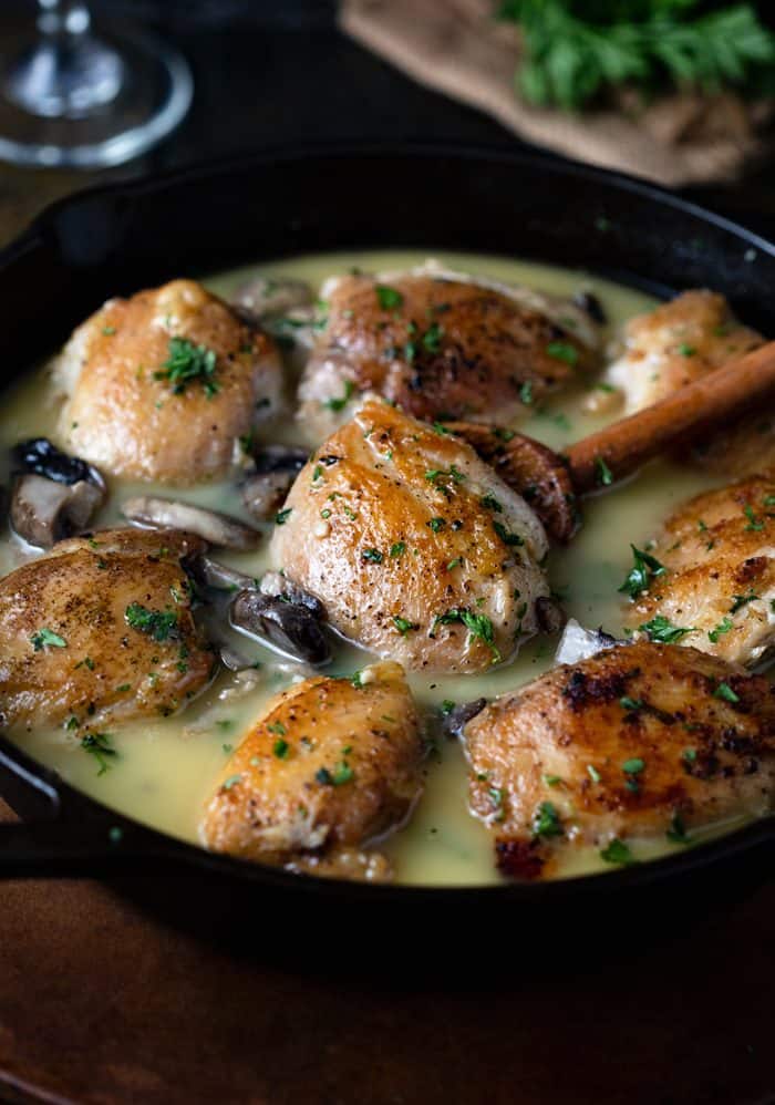 A wooden spoon lifting up a baked chicken thigh from a large cast iron skillet filled with chicken and wine sauce.