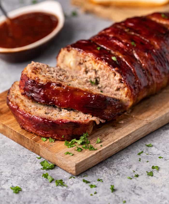 Meatloaf sliced on a wooden cutting board with fresh parsley sprinkled on top and homemade ketchup sauce in background