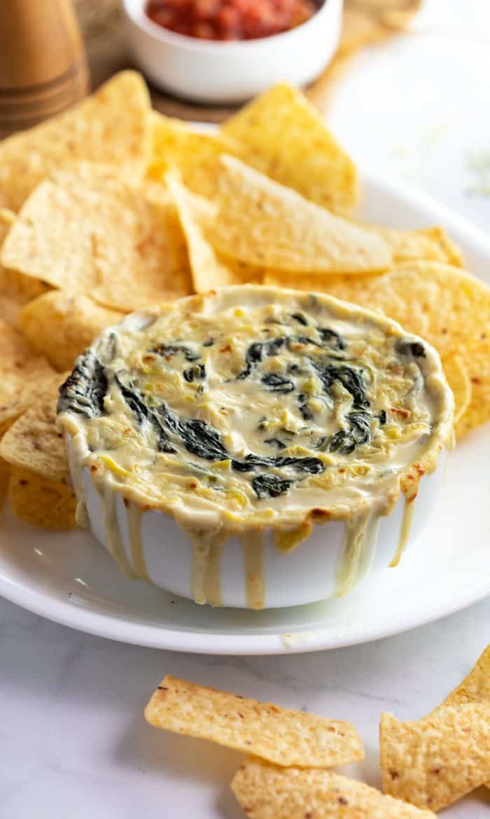 White bowl overflowing with spinach artichoke dip and corn tortillas on white plate