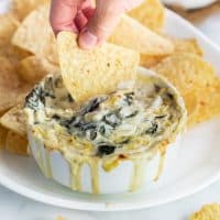 A hand dipping a tortilla chip into a white bowl of Applebees Spinach and Artichoke Dip.