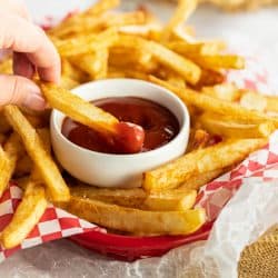A hand dipping homemade french fries into a white cup of ketchup.