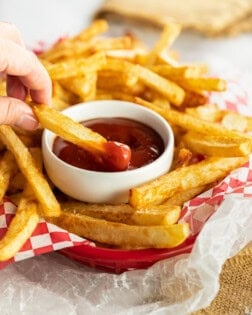 A hand dipping homemade french fries into a white cup of ketchup.