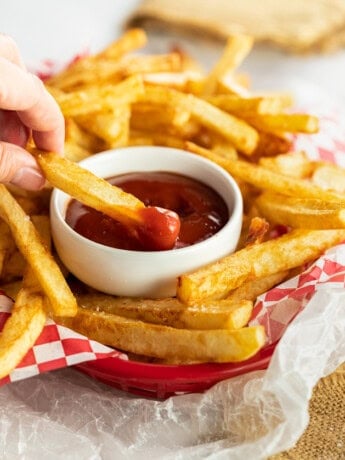 A hand dipping homemade french fries into a white cup of ketchup.