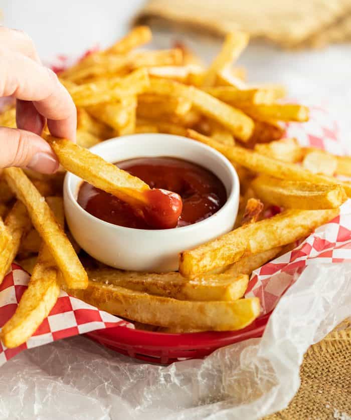 Hand dipping crispy homemade french fry into a white bowl of ketchup inside a basket of fries