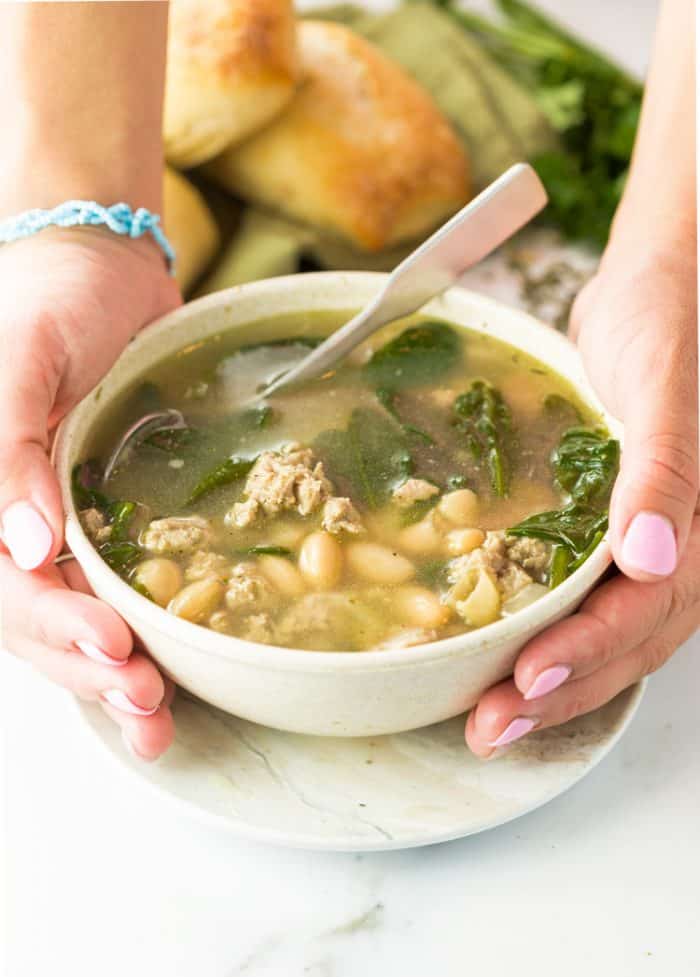 Two hands holding a white bowl filled with warm soup with ground turkey, white beans, and spinach in broth.