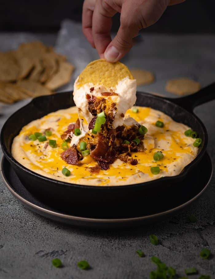 A hand dipping a tortilla chip into bacon ranch cheese dip in a black cast iron skillet.