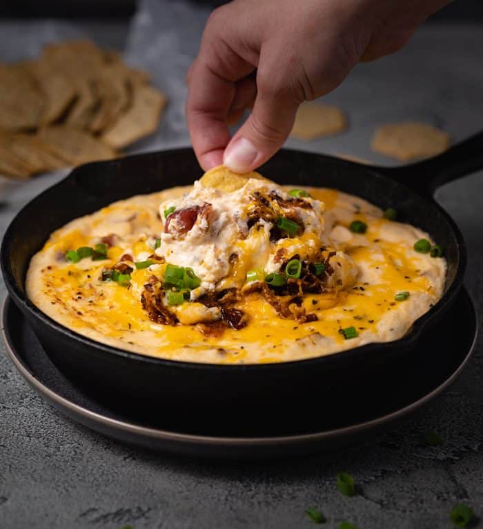 A black cast iron skillet filled with bacon ranch cheese dip with a hand dipping a chip into it.