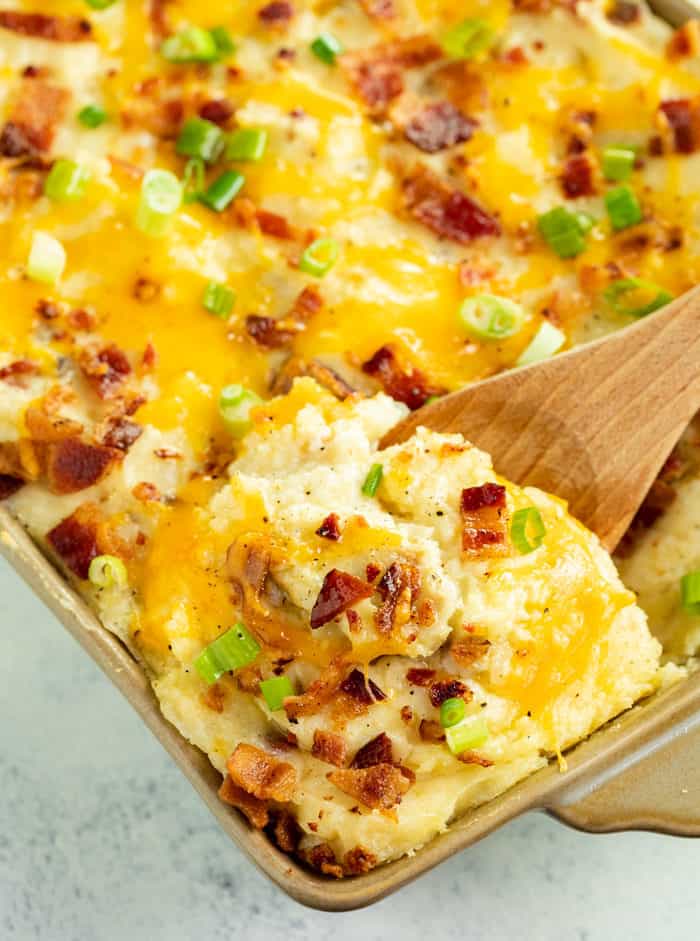 A wooden spoon scooping out twice baked potato casserole topped with melted cheese, bacon, and green onions.
