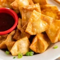 A homemade Crab Rangoon on a white plate with chives and sweet and sour sauce.