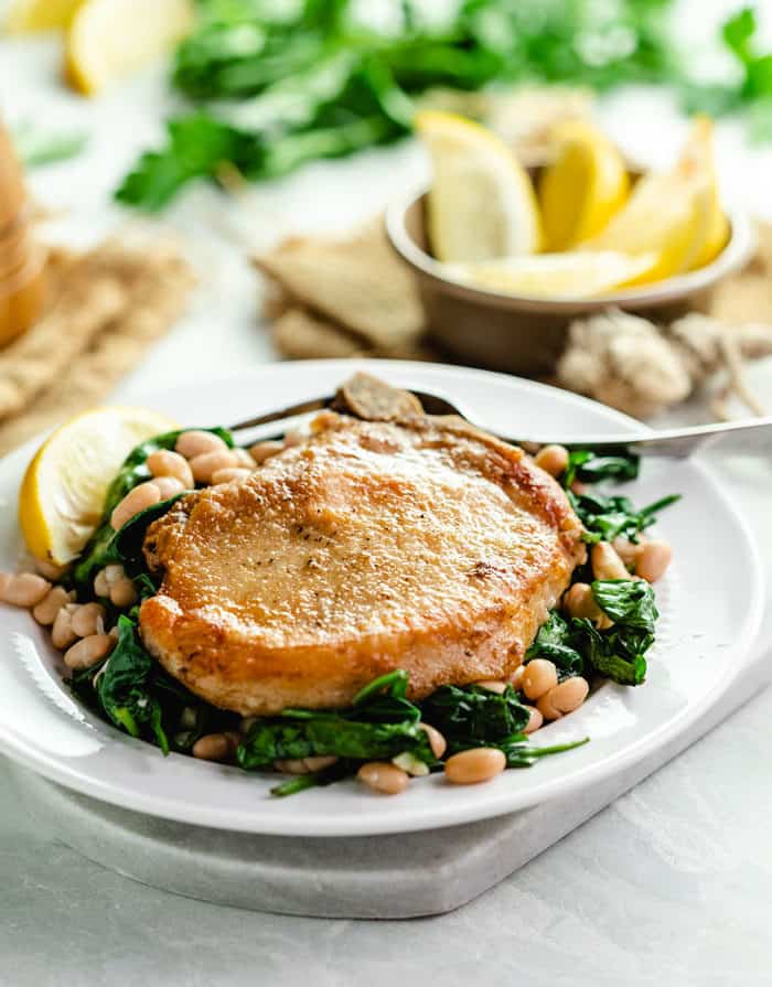 Pan fried pork chops on a bed of spinach and white beans on a white plate with lemon wedges in background.