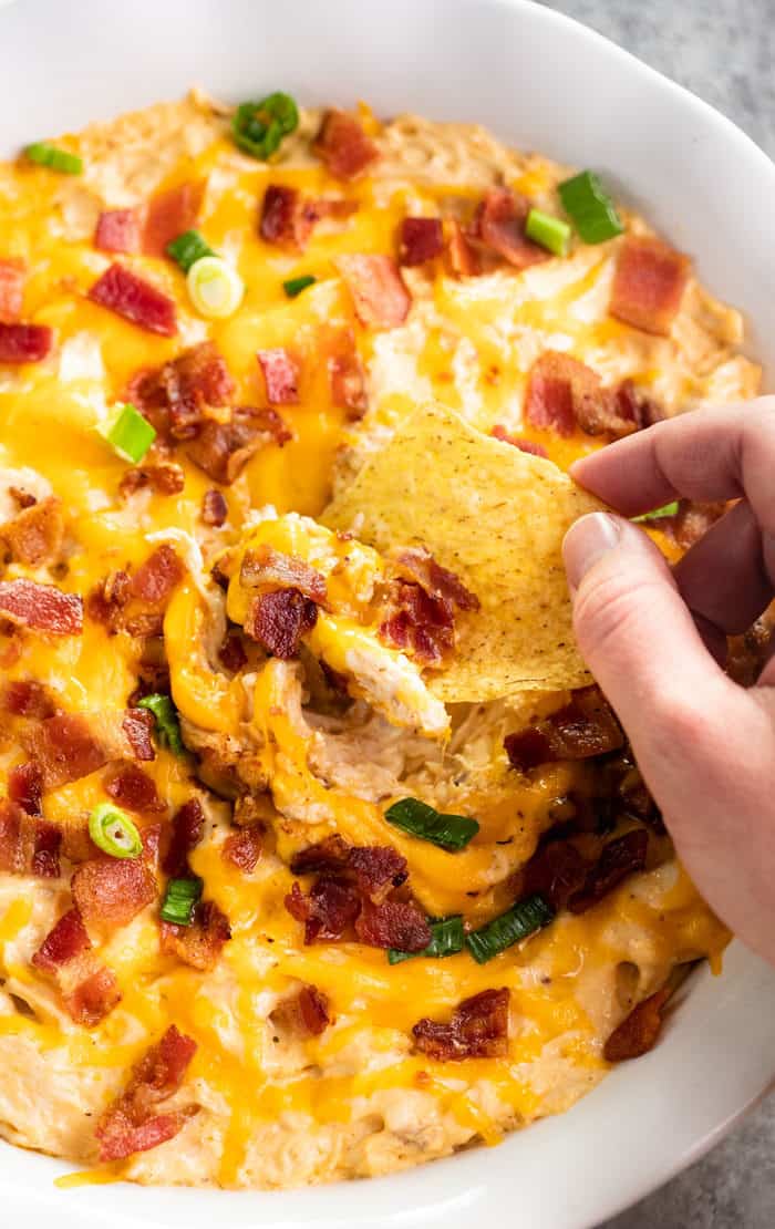 Close up image of hand holding tortilla chip and dipping into white bowl of chicken ranch bacon dip