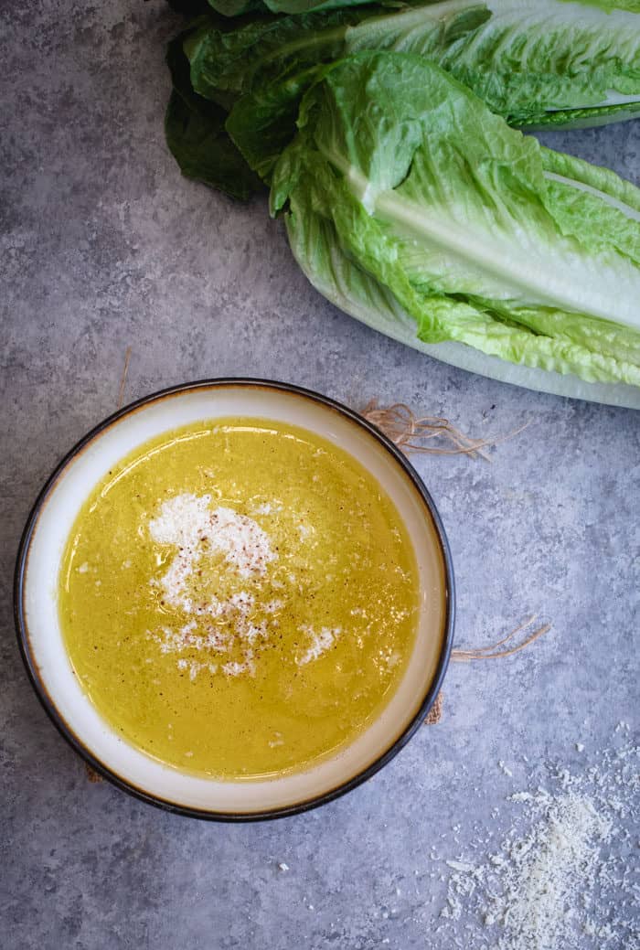 A bowl filled with homemade caesar salad dressing with Parmesan cheese sprinkle on top and romaine lettuce next to it.