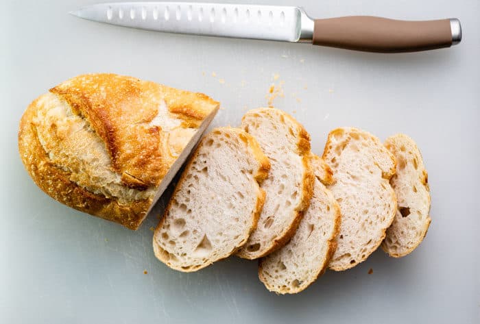 loaf of sourdough bread half sliced on a white cutting board with a knife.