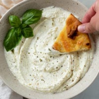 A hand dipping bread into a bowl of Feta Dip garnished with basil.