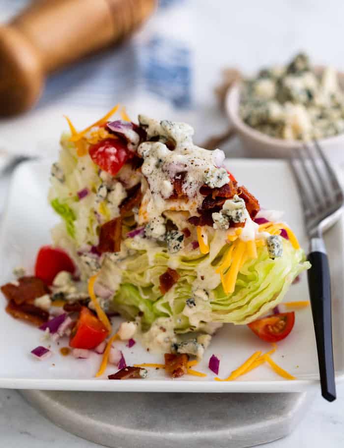 Wedge salad on a white plate topped with blue cheese, bacon, tomatoes, and cheddar.